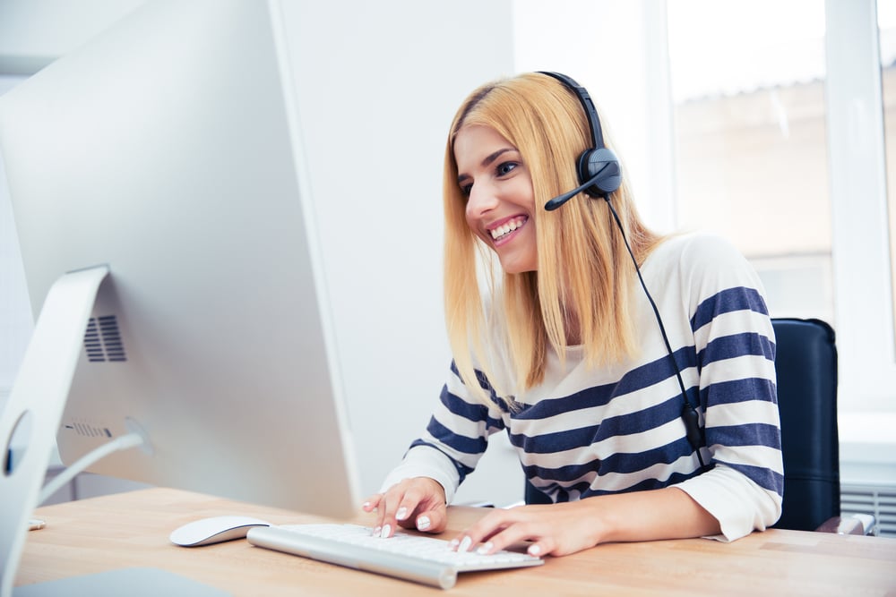 Cheerful young female operator with headset using desktop computer in office